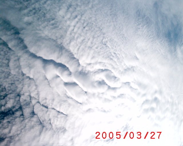 Aerial view of thick white cloud formations with layered, wave-like patterns against gray sky, dated 2005/03/27