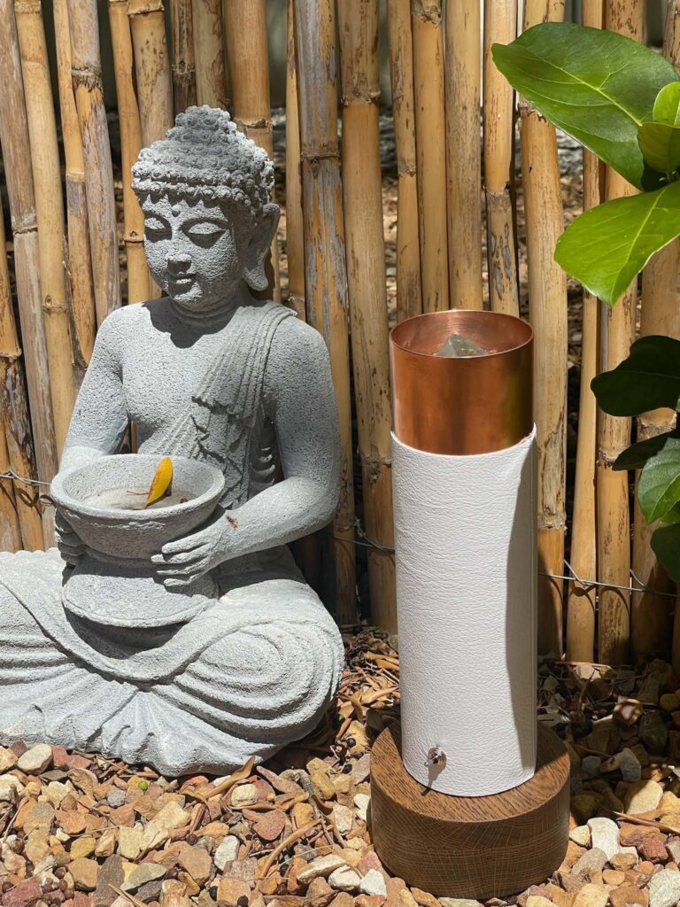 Steinerne Buddha-Statue mit Schale und gelbem Objekt neben weiß-kupfernem zylindrischem Gerät auf steinigem Boden mit Bambuszaun