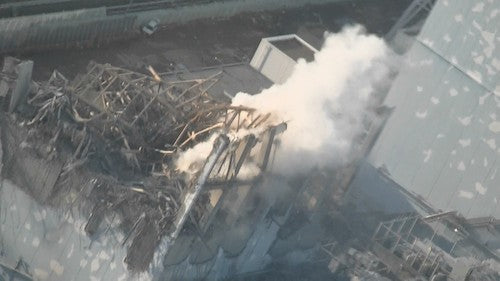 Aerial view of a damaged building with collapsed roof structure, debris, and smoke or dust rising from the wreckage.