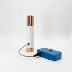 Tube cylindrique blanc avec extrémités métalliques cuivrées relié par câble gris à boîtier de contrôle bleu rectangulaire