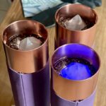 Trois tasses métalliques cuivrées avec étuis en cuir violet, contenant liquide foncé et glaçons, une éclairée en bleu