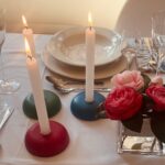Drei brennende weiße Kerzen in bunten runden orgonite-Haltern (rot, grün, blau) auf weißer Tischdecke mit rosa Blüten