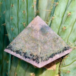 orgonite rosa en forma de pirámide con virutas metálicas y materia vegetal, colocado sobre cactus verde con espinas visibles