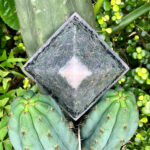 Pirámide orgonite cuadrada con superficie marmoleada verde oscuro y cristal rosa central, colocada sobre cactus verde rodeado