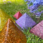 Bunte orgonite Pyramiden auf Gras mit lila Blüten - bernstein, gelbe, rote, lila und rosa Harzpyramiden mit Metallflocken