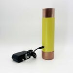 Appareil cylindrique jaune avec extrémités métalliques cuivrées et adaptateur secteur noir avec cordon sur fond blanc