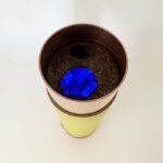 Récipient cylindrique avec base jaune, section médiane beige, rebord supérieur marron. Contient matière fibreuse sombre et pierre bleue.