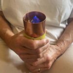 Mains tenant cylindre en cuivre avec bande verte autour de la base, contenant objet pyramidal bleu à l'intérieur