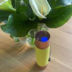 Appareil cylindrique avec bande textile jaune et extrémités brunes, cristal bleu visible à l'intérieur, près d'une fleur blanche
