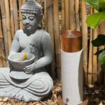 Weißer zylindrischer orgonite Stab mit Kupferspitze auf Holzsockel, neben grauer Buddha-Statue mit gelber Blüte, Bambuszaun