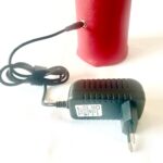 Appareil orgonite cylindrique rouge avec connecteur doré et câble noir relié à adaptateur secteur noir sur surface blanche