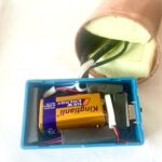 Boîtier plastique bleu avec batterie 9V, composants électroniques noirs et fils. Dispositif orgonite rond cuivré au-dessus.