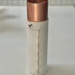 Étui cylindrique en cuir blanc avec coupelle métallique cuivrée au sommet, fermeture à bouton-pression et zip, debout