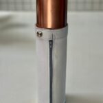 Étui cylindrique en cuir blanc avec haut métallique cuivré, fermeture éclair verticale, bouton-pression, debout sur surface