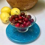 Blaue runde Ladeplatte mit klarer Glasschale voller roter Kirschen, drei gelben Zitronen und geflochtenem Korb auf weißer Fläche