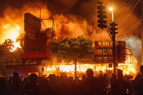 Night scene showing buildings on fire with smoke, crowds of people silhouetted in foreground, street signs visible