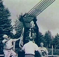 Black and white photo of large cylindrical tubes or pipes mounted at an angle, with people standing nearby for scale