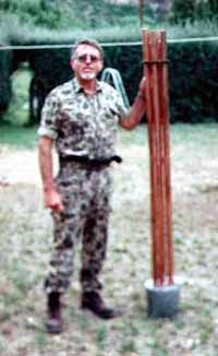 Person in camouflage uniform standing outdoors holding tall reddish wooden stakes or poles, dirt ground visible