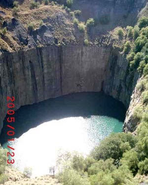 Aerial view of deep circular crater filled with turquoise water, surrounded by rocky cliffs and green vegetation