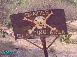 Weathered danger sign with skull and crossbones warning of blasting area, mounted on post in arid desert terrain with sparse vegetation
