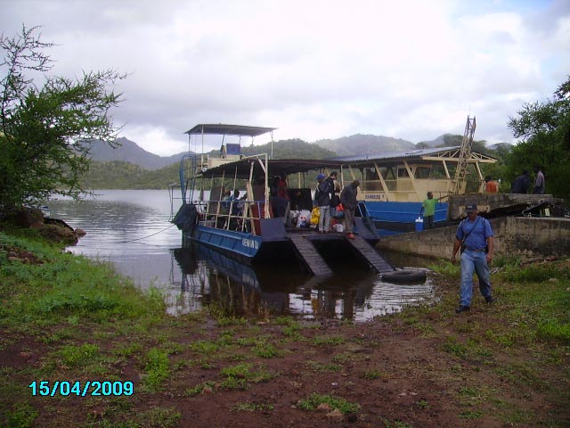 Boat docked at muddy shoreline with passengers aboard, man standing on shore, lake and hills in background, overcast sky
