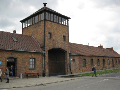 Brick building with watchtower, arched entrance, red tile roofs under cloudy sky. People visible near entrance.