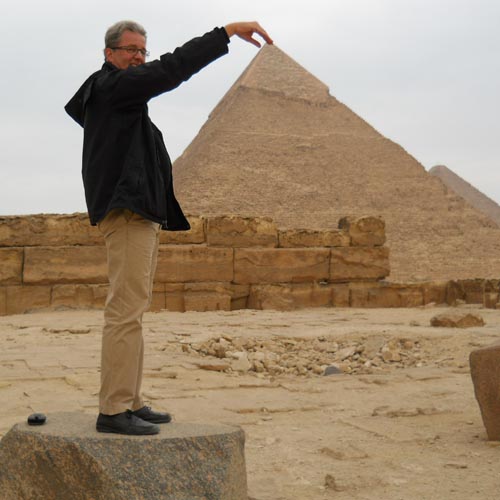 Man in dark jacket posing with hand positioned to appear touching pyramid tip, standing on stone block at Giza pyramids