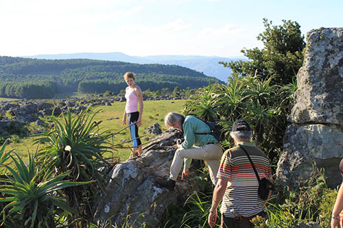People on rocky outcrop with vegetation, overlooking valley with village and forested hills under partly cloudy sky