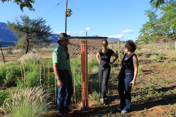 Three people stand near a tall metal pole with orgonite device in rural terrain with mountains, trees, and blue sky