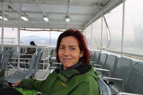 Woman in green jacket sitting on ferry deck with plastic chairs, mountains and water visible in background