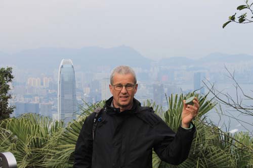 Man with glasses in dark jacket standing outdoors with city skyline and mountains in background, vegetation in foreground