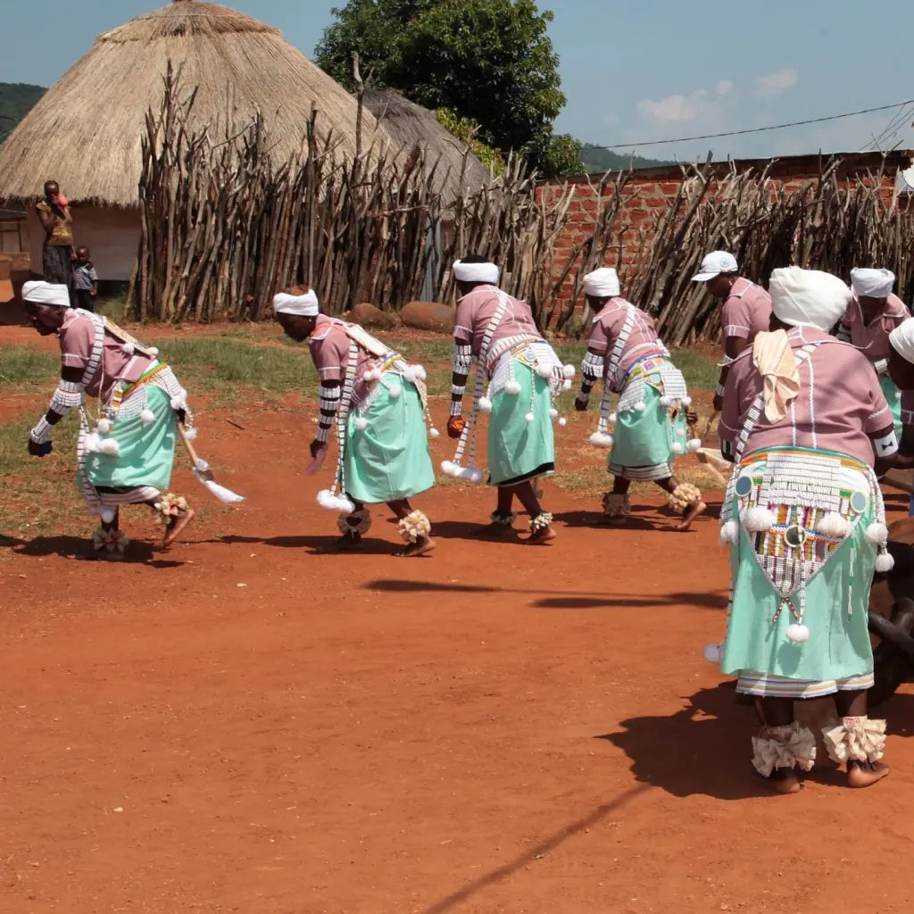 Traditional dancers in colorful robes and white head wraps perform on red earth, with thatched huts and village buildings visible