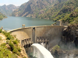 Large concrete dam with water spillway releasing white water, surrounded by forested mountains and a reservoir lake