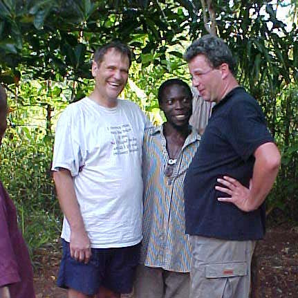 Three men pose together outdoors under leafy trees, two wearing casual shirts and one in striped clothing, smiling at camera