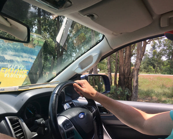 Driver's view from inside vehicle showing hand on steering wheel, rural road with trees and hills visible through windshield
