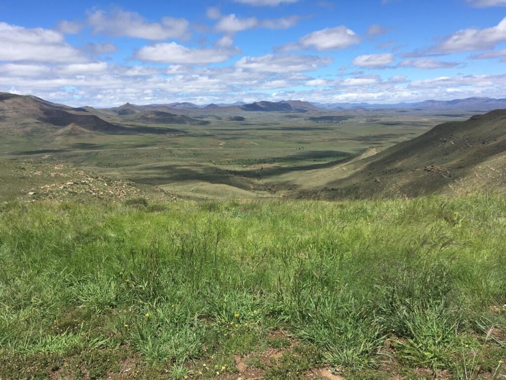 Rolling green hills and distant mountains under blue sky with white clouds. Green grass in foreground, arid landscape beyond.