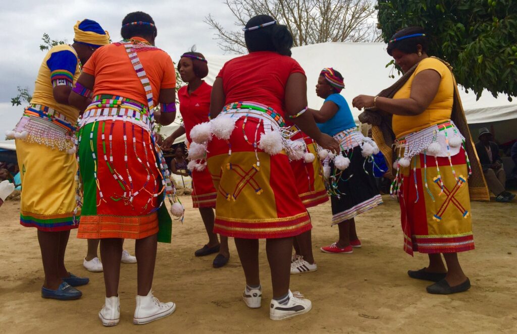Group of people in colorful traditional African clothing with beadwork and white pom-poms dancing outdoors on sandy ground