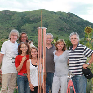 Group of seven people posing outdoors with tall copper-colored cylindrical device, green hills in background